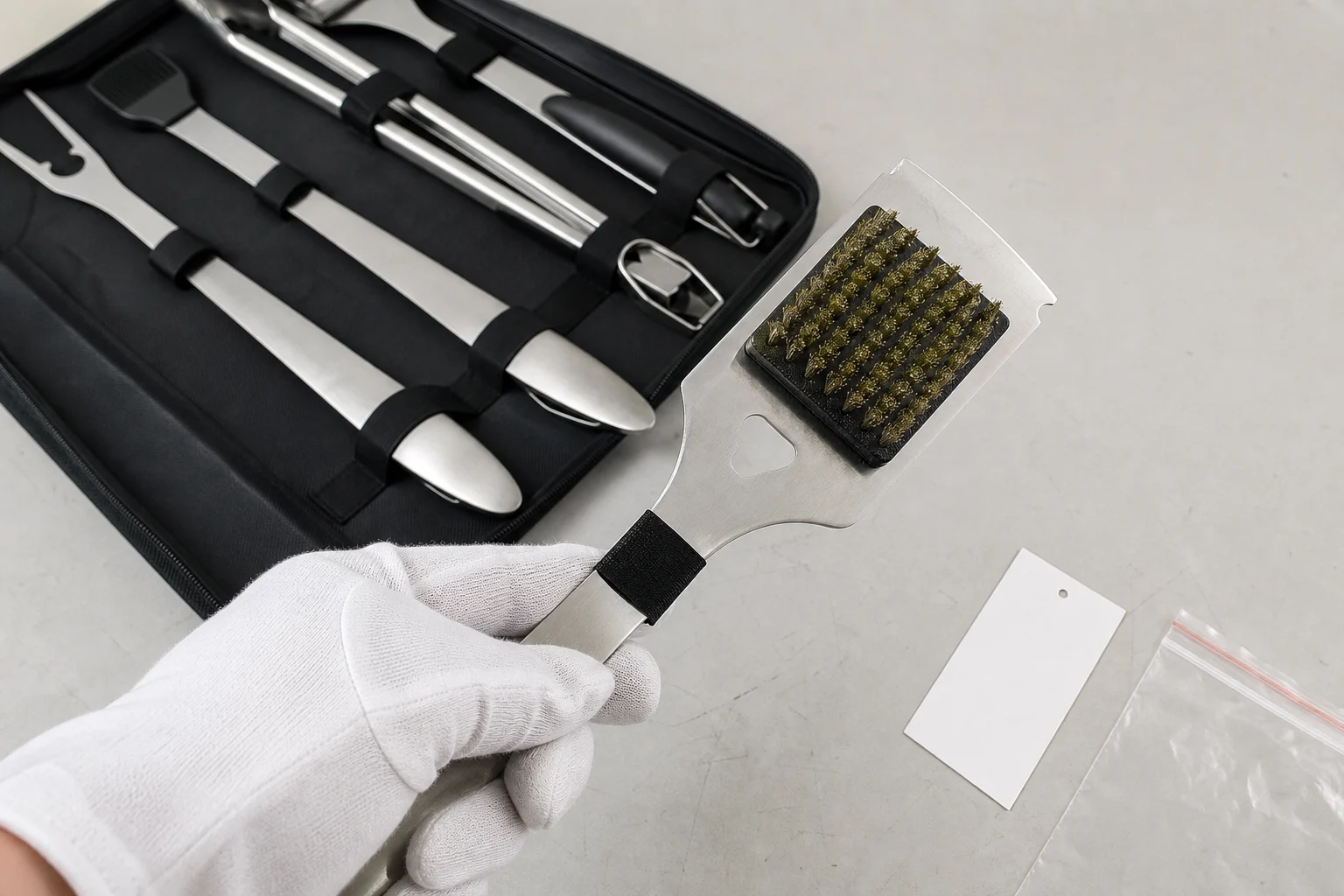 Gloved inspection of a clean Grillive grill cleaning brush beside an opened BBQ tool set and blank label card